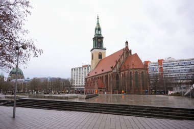 Yeşil bakır saat kulesi olan tarihi bir tuğla kilise, modern binalar, kubbeli bir katedral ve şehir manzarasıyla çevrili ıslak bir şehir meydanında duruyor.. 