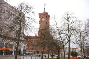 Berlin, Almanya 02.13.2025 Büyük kırmızı tuğlalı saat kulesi bulutlu bir günde modern binalar, dükkanlar ve yayalarla çevrili hareketli bir şehir manzarasında yapraksız ağaçlar arasında duruyor..