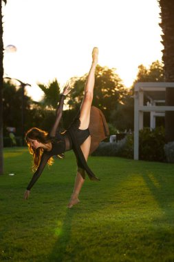 Siyah elbiseli zarif bir balerin yemyeşil bir çimenlikte altın gün batımı ışığında uzun bir bacak uzantısı yapar..