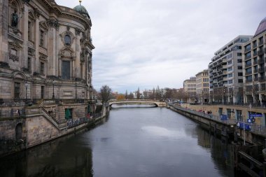 Berlin 'deki Spree Nehri' nin barışçıl bir manzarası, tarihi ve modern mimari çerçeveli, bulutlu bir gökyüzünün altında bir köprü..