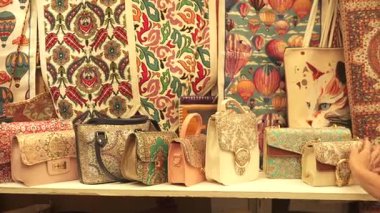 Bir kadın süslü el çantaları ve nakışlarla dolu, karmaşık desenler, canlı renkler ve geleneksel kültür işçiliği içeren bir butik market tezgahını keşfediyor..