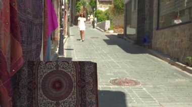 Beyaz elbiseli bir kadın, tarihi bir bölgede güneşli bir kaldırım taşı caddesinde renkli halılarla süslenmiş bir şekilde geziniyor. Antalya Türkiye kenti