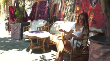 Beyaz elbiseli çıplak ayaklı bir kadın bambu bir sandalyede oturur, kabuk halhalhalını düzeltir, örülmüş halılar, kırsal dekorlar ve boho atmosferinde yumuşak benekli güneş ışığıyla çevrilidir.