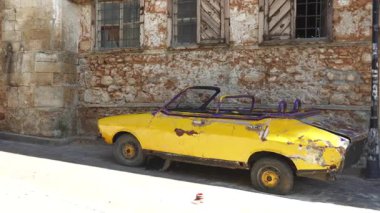 Paslı, sarı, üstü açılabilir bir araba, tahtadan pencereli yıpranmış taş bir duvarın yanında hasarlı ve unutulmuş bir şekilde duruyor. Nostaljik ve antika bir kentsel çürüme sahnesi yaratıyor..
