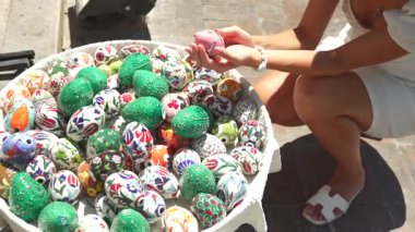 Beyaz elbiseli bir kadın açık hava pazarında elde boyanmış seramik yumurtaya hayran. Etrafı canlı el yapımı hediyelik eşyalar, el sanatları ve kültürel hazinelerle çevrili.