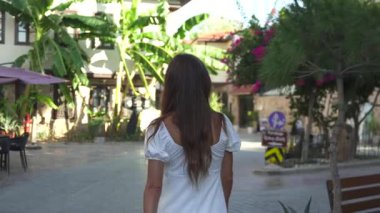 Etrafında taş binalar, çiçek açan begonviller ve açık hava kafeleri olan sevimli bir kasaba meydanında yürüyen genç bir kadın. Antalya Şehri 