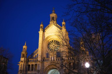 Brüksel 'deki görkemli Aziz Catherine Kilisesi mavi saat boyunca aydınlandı, önplanda karmaşık mimari ve yapraksız ağaçlarla.