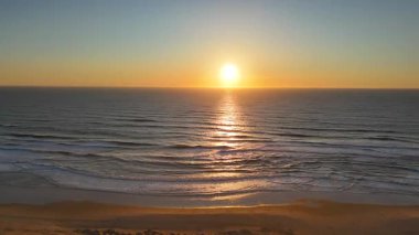 İnsansız hava aracı, okyanusun üzerinde yumuşak dalgalar ve kumlu kumsal ile altın bir gün batımı yakalar. Huzurlu, rüya gibi bir sahne yaratır..