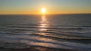 İnsansız hava aracı sakin okyanus dalgaları üzerinde sakin bir gün batımını yakalar, sakin suyun üzerinden sıcak akşam ışığında boş bir sahile doğru uçar.. 