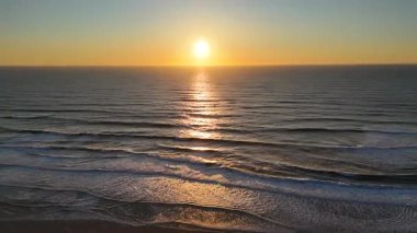 Sakin okyanus dalgaları kumlu sahil boyunca sıcak gün batımını yansıtarak altın alacakaranlıkta sakin ve atmosferik bir kıyı manzarası oluşturur.. 