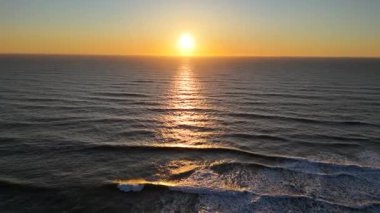 Okyanusun yukarısındaki altın bir günbatımının hava görüntüleri. Kameralar dalgaların üzerinden sakin, boş bir kumsala doğru süzülüyor.