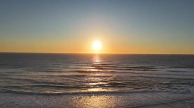 Okyanus dalgalarının üzerine sıcak bir ışık tutarak, huzurlu ve doğal güzellikte boş bir kumsalı gözler önüne seren, altın günbatımının hava görüntüsü. 