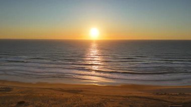 İnsansız hava aracı okyanusun üzerinde altın gün batımını yakalar, sakin dalgaların üzerinde süzülür ve sıcak akşam ışığı altında ıssız kumlu bir plaj.