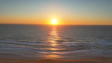 İnsansız hava aracı, gün batımında yumuşak ve huzurlu bir sahil şeridine yuvarlanan köpüklü dalgalarla dokulu okyanus dalgalarını yansıtan altın gün batımını yakalar..