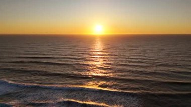 Sakin okyanus sularının üzerinde geniş açılı drone görüntüsü yumuşak dalga katmanları ve deniz boyunca uzanan parlayan yansıması barışçıl, sinematik bir atmosferde..