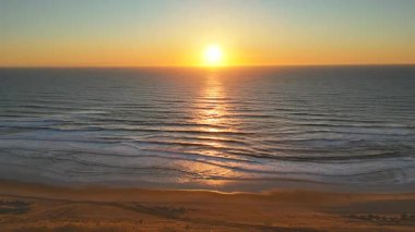 Sakin okyanusun üzerinde, ışığı yansıtan dalgalarla ve önplanda barışçıl kumlu bir plajla altın günbatımının havadan çekilmiş görüntüsü. Alacakaranlıkta sakin, manzaralı bir sahil anı.