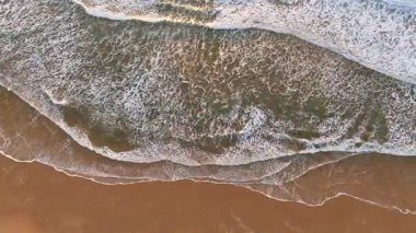 Yukarıdan okyanus sörf modellerinin havadan görünüşü, köpüklü dalgalar, sakin ve sıcak kumlu bir plajın üzerinde süzülüyor günbatımında huzurlu ve dokulu bir sahil anı.. 