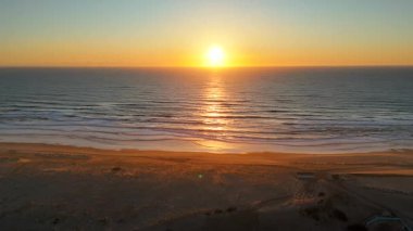 Sahil köyü Loncau 'nun altın gün batımındaki hava aracı görüntüsü, ışıldayan okyanus ufkunu, yumuşak dalgaları ve ılık akşam ışığında yıkanan huzurlu plajı gösteriyor. Seyahat veya editoryal kullanım için ideal. 