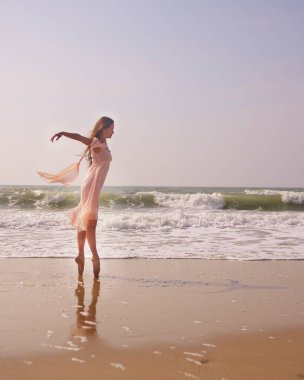 Zarif bir balerin, yumuşak, pembe bir elbise giyerek okyanusun kenarında dikiliyor. Huzurlu deniz kenarı, sanat ve doğa arasındaki zarafeti, hareketi ve uyumu vurgular.. 