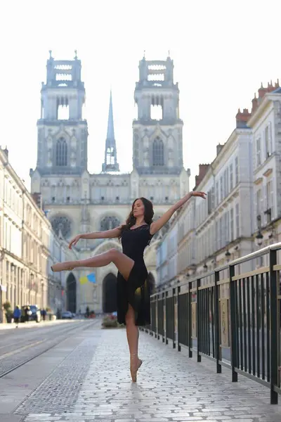 Siyah elbiseli balerin, arka planda katedral kuleleri yükselen kaldırımlı bir sokakta zarif bir şekilde dengede duruyor.. 