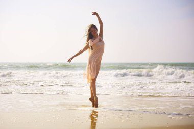 Arkasından dalgalar yuvarlanırken bir balerin ıslak kumun üzerinde dikiliyor. Hafif bir elbise içinde, sakin bir sahil sabahının narin ışığı altında zarif bir poz verir.. 