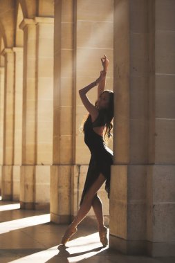 Siyah elbiseli zarif balerin, klasik bir taş atari salonunda sıcak güneş ışığıyla yıkanmış bir sütuna karşı noktasında gösteri yapıyor.. 