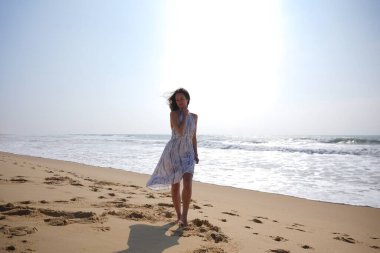 Genç bir kadın kumlu sahilde çıplak ayakla yürüyor açık bir elbiseyle, arkasında okyanus dalgaları ve parlak güneş ışığı ile.. 