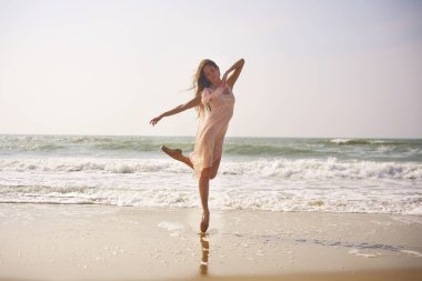 Zarif bir balerin, kumlu bir plajda düz bir elbise giyerek dengede durur. Anlamlı duruşu ve arkasındaki yumuşak okyanus dalgaları doğadaki zarafeti, özgürlüğü ve hareketi çağrıştırıyor.. 