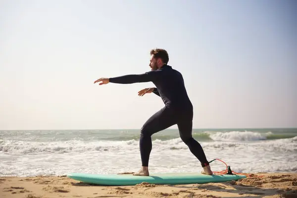 Dalgıç kıyafetli bir adam kumlu bir plajda sörf tahtası üzerinde sörf yapıyor. Dengeye ve forma odaklanıyor.. 