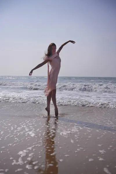 Narin pembe elbiseli bir balerin okyanusun kenarında ayak parmaklarının üzerinde yükseliyor. Kolları zarif bir şekilde hareket ediyor yansıması yumuşak dalgalar ve açık gökyüzü altında ıslak kumda uzanırken.. 