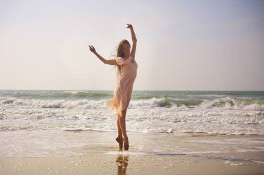 Bir balerin, şeftali rengi bir elbise giyerek kumsalda zarif bir poz verir. Kollarını gökyüzüne kaldırırken, dalgalar arkasından süzülüyor zarafet, özgürlük ve hareketi yakalıyor.