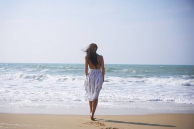 Genç bir kadın dalgalı bir yaz elbisesiyle sahilde tek başına yürüyor ve denize yaklaşırken kumların üzerinde ayak izleri bırakıyor. Suyun kenarında huzurlu bir yalnızlık ve özgürlük anı.. 