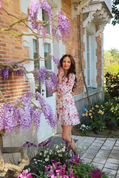 Gül desenli elbisesiyle çekici bir kadın bahar çiçekleriyle çevrili bir tuğla evin önünde çiçek açan mor salkımın yanında duruyor.. 