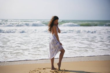 Akan bir elbisesi olan genç bir kadın sahilde yalınayak yürüyor, arkasında dalgalarla yavaşça kuma dokunuyor.. 