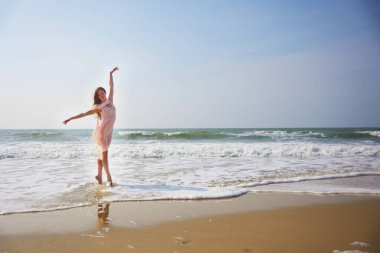 Zarif bir balerin su kenarında pembe bir elbise giyerek dans eder. Zarif duruşu köpüklü dalgalar ve açık mavi gökyüzü ile çok güzel zıtlaşıyor..