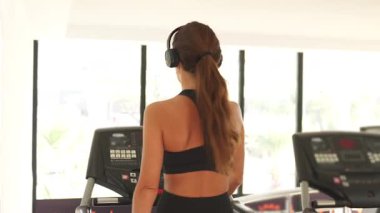Uzun saçlı kadın, iyi ışıklandırılmış spor salonunda koşu bandında yürüyor, kablosuz kulaklık takıyor ve hafif kardiyo ve sağlığa odaklanıyor..