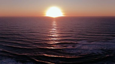 Hava aracı görüntüsü, güneşin sakin bir okyanusun üzerinde battığını gösteriyor. Yumuşak pembe ve turuncu yansımaları, dalgalı dalgalar boyunca yayıyor..