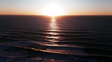 Altın güneş ışığı, ufukta güneş batarken yumuşak okyanus dalgaları boyunca yankılanır, sakin bir hava aracı görüntüsüyle yakalanır.