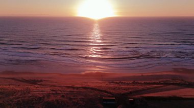 Drone, sakin okyanus dalgaları üzerinde güneşin batışını yakalar. Suyun üzerinde ve kumlu sahilde yumuşak akşam ışığında altın-pembe bir yansıma oluşturur..