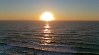 Parlak bir günbatımının havadan görünüşü narin okyanus dalgaları üzerinde altın yansımalar oluşturarak huzurlu ve geniş bir deniz manzarası yaratıyor..