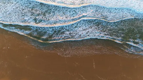 Yukardan aşağıya doğru ıslak kumda yuvarlanan köpüklü mavi dalgaları yakalar, kıyı şeridi boyunca doğal çizgiler, yansımalar ve dokular oluşturur..
