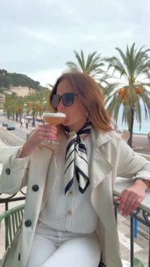 Güneş gözlüklü ve trençkotlu zarif kadın, Fransa 'nın Nice kentinde palmiye ağaçları ve okyanus manzaralı bir terasta kokteyl içiyor.. 