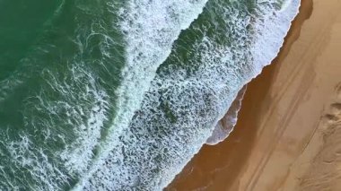 Yan taraftaki drone görüntüsü kumlu sahile vuran köpüklü dalgalar gösteriyor, lastik izleri ve manzaralı kıyı şeridi boyunca detaylı kum desenleri var..