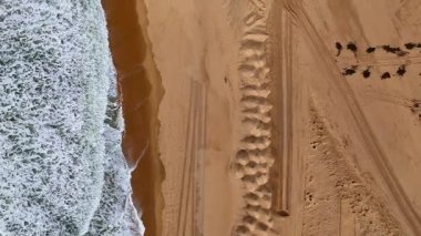 Havadan çekilen drone, lastik izleri, su hatları ve doğal dokularla işaretlenmiş kumlu bir plaja vuran ayrıntılı köpüklü dalgaları yakalar..
