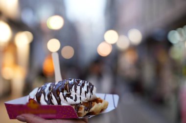 Arka planda bulanık şehir ışıklarıyla çırpılmış krema ve çikolata sosuyla kaplı Belçika waffle 'ını tutan bir elin yakın çekimi..