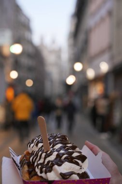 Çırpılmış krema ve çikolatayla kaplı Belçika waffle 'ını kaldırımları ve sokak lambalarını bulanık bir şekilde tutan bir elin yakın çekimi..