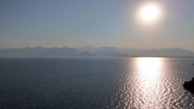 Hala insansız hava aracı görüntüsü parıldayan denizi, uzak dağ siluetlerini ve parlak batan güneşi ufuk boyunca altın ışığı yansıtıyor..