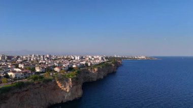 İnsansız hava aracı rotasyonu, yüksek binalar, okyanus kayalıkları ve uzak dağların üzerinde parlayan günbatımının yer aldığı kıyı şehri manzarasıyla sona eriyor.. 