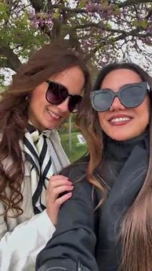 Şık kıyafetler ve güneş gözlüğü takan neşeli kadınlar bahar çiçekleri ve ağaçlarla dolu güzel bir parkta selfie çekiyorlar. Paris, Fransa 04.27.2025 