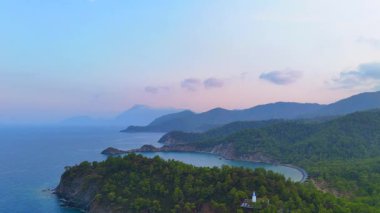İnsansız hava aracı panoraması, sakin bir Akdeniz kıyı şeridi boyunca güneşin doğuşuyla yıkanmış dramatik orman uçurumlarını, tenha bir körfezi ve katmanlı dağları gösteriyor.. 